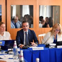 Uomini e donne in riunione di lavoro, discussione al tavolo con documenti e bottiglie d’acqua, sullo sfondo specchi che riflettono i partecipanti.