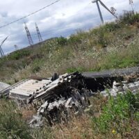 Veicolo sfasciato su un terreno erboso, alberi bassi e linee elettriche in secondo piano, cielo nuvoloso.
