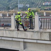Tre vigili del fuoco in tuta fluorescente blu/verde sul ponte di una strada, con attrezzature, allontanano con cautela un corpo estraneo dall’orlo.