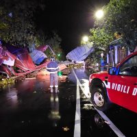 Auto della viabilità d’emergenza e vigili del fuoco alla notte, sirene non in funzione, strada allagata e tende rosse su entrata improvvisata.