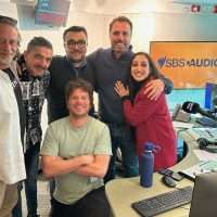Gruppo di sei persone in ufficio radiotelevisivo SBS Audio, sorridenti, pose informali davanti a postazioni con computer e microfoni.