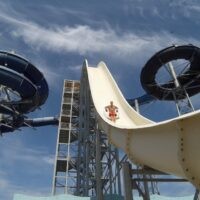 Scivolo alto in parco divertimenti con tubi scuri e piattaforma, cielo azzurro, elementi di grisaglia e logo centrale.