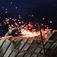 Operai su una passerella metallic a bordo di un tetto, scintille rosse esplodono nell’aria durante lavori vicino al buio della notte.