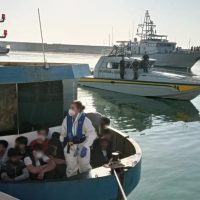 Gruppo di migranti su una piccola barca blu nelle acque tranquille, con una nave al largo e un ufficiale in uniforme al centro.