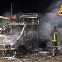 Van incendiata e fumo sullo sfondo, vigili del fuoco in azione durante l’intervento di spegnimento.
