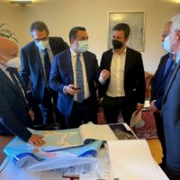 Gruppo di sei uomini in abiti formali con mascherine, discutono attorno a una tavola con mappe e documenti.