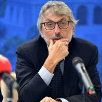 Imprenditore elegante con occhiali, barba e mani sul mento durante una conferenza, microfono davanti, sfondo blu sfumato.