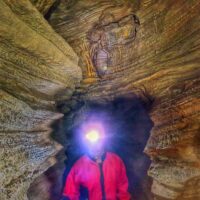 Escursionista in una grotta, caschetto con torcia accesa illumina pareti rocciose e passaggi stretti.