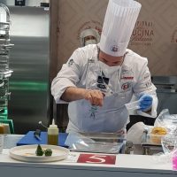 Chef in abiti bianchi con cappello, impegnato a decorare un piatto su tavolo da cucina professionale, utensili e foglia verde accanto.