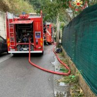 Vigili del fuoco a bordo di autopompa rossa, tubo flessibile disteso lungo la strada, operazione di spegnimento in corso vicino a recinzione verde.