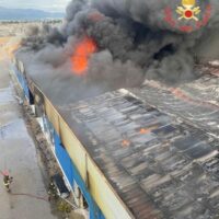 Incendio su un tetto di un edificio industriale, colonne di fumo nero si alzano e fuoco visibile tra le tegole, persiste pericolo.