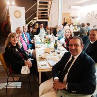 Gruppo di persone a tavola in una riunione o pranzo aziendale, sorridenti, ambiente luminoso e moderno, con cibo e vetri sul tavolo.