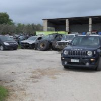 Parcheggio all'aperto con jeep e SUV, veicoli sportivi su superficie sterrata, sfondo capannoni industriali e cielo nuvoloso.