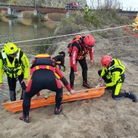 Operatori di soccorso in tuta nera e giubbotto ad alta visibilità lavorano su una zattera arancione vicino a un fiume, sorveglianza e coordinazione in emergenza.