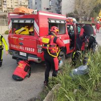Vigili del fuoco in azione vicino a un’auto ferita, zaini e attrezzature sulla strada, veicolo di soccorso rosso sullo sfondo.