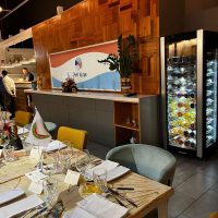 Interno ristorante elegante con tavoli pronti, bicchieri e posate, scaffali in legno, atmosfera accogliente e frigorifero pieno di bevande sullo sfondo.