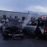 Automobili distrutte, fiamme e fumo si alzano da due veicoli incendiati in un'area industriale al crepuscolo.