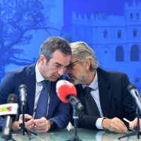 Due uomini al tavolo conferenza, uno con microfono rosso, intento a discutere davanti a una facciata blu di edificio storico.