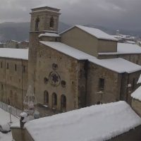 Chiesa romanica di pietra con campanile, coperta di neve, vista panoramica su tetti innevati e sfondo nuvoloso