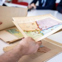 Mano tiene una carta de mapa o documento antiguo sobre una mesa, con varias envelopes apiladas, en un entorno de oficina.