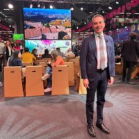 Uomo in abito scuro posa sorridente in sala conferenze, sfondo con grandi schermi e pubblico seduto, atmosfera vivace e luminosa.