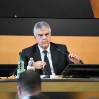 Uomo in abito in sala conferenze, seduto al tavolo con microfono e bottiglia d'acqua, sfondo neutro, attenzione al discorso.