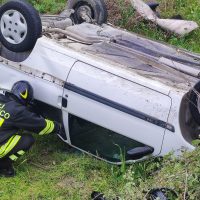 Auto ribaltata su fianco in prato verde, passeggero soccorritore all’ingresso del veicolo, scenario di incidente stradale.