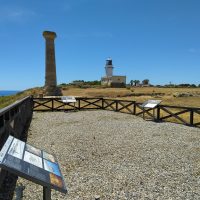 Vista panoramica di resti storici in pietra e una torre sulla costa, con cielo azzurro e sentiero ghiaioso recintato.
