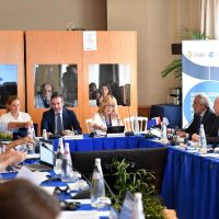 Foto di una riunione di business: partecipanti seduti attorno a un tavolo, discussione animata, bottiglie d'acqua e documenti, ambiente conferenze.