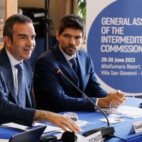 Due uomini in giacca e cravatta parlano a una conferenza; sfondo con bandiere e testo, atmosfera professionale e formale.