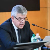 Uomo con capelli grigi e occhiali, seduto a un tavolo, parla al microfono durante una conferenza o incontro ufficiale, con bottiglie d'acqua vicino, sfondo neutro in ambito professionale.