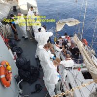 Uomini in tuta bianca su un'imbarcazione da ricerca, presenti su una nave da soccorso in mare aperto, contesto operativo di assistenza.