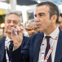 Uomo in abito che annusa un bicchiere di liquore dorato in un evento, con altri partecipanti sullo sfondo.