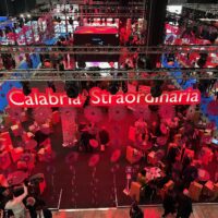 Vista dall’alto su una fiera o evento aeree; insegna luminosa rossa Calabra Strar...; folla, stands e attività in uno spazio espositivo affollato.