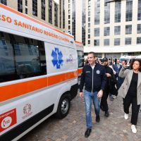 Ambulanza in primo piano, operatore sanitario e cittadini camminano nel cortile di un ospedale con edifici moderni sullo sfondo.