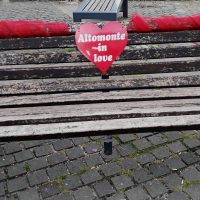 Cuore rosso con scritta Automenzione in love appeso a una panchina di legno, su ciottoli di pavimentazione.