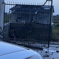 Sedia e rottami sparsi dietro una recinzione metallica vicino a una struttura danneggiata, tracce di lesione a terra e veicolo in primo piano.