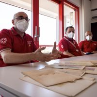 Tre operatori sanitari con mascherine e camici rossi, seduti a un tavolo, esaminano documenti medici su una scrivania.