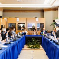 Riunione formale in sala conferenze con tavolo a forma di U, partecipanti seduti e discussione animata, ambiente professionale.