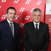 Due uomini in abiti formali sorridono davanti a una parete rossa con logo bianco, in un contesto ufficiale o di conferenza.
