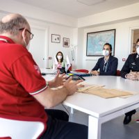 Gruppo di persone, inclusa una persona anziana in camicia rossa, sedute intorno a un tavolo bianco in sala riunioni: discussione, documenti e mascherine.