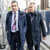 Due uomini in abiti formali e giubbotti scuri passeggiano in una zona affollata, con badge appesi al collo e strutture metalliche sullo sfondo.