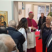 Un vescovo in abito liturgico benedice un gruppo di persone in una sala espositiva, tra dipinti e una cornice rossa del percorso.