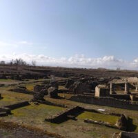 Veduta panoramica di rovine archeologiche all’aperto con cappelli di pietra e mura parzialmente conservate, cielo azzurro e nuvole leggere.