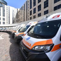 Furgoni bianchi con strisce arancioni, linea di ambulanze o mezzi di soccorso parcheggiati in fila su strada lastricata, fronte urbano.
