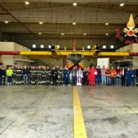 Gruppo di persone in uniforme allineate in un hangar industriale, con bandiere e insegne sullo sfondo, illuminazione diffusa.