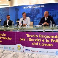 Tribuna in sala conferenze; tre uomini discutono sul tavolo, sfondo blu con testo Regione Calabria, evento sui servizi e lavoro.