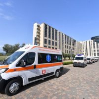 Ambulanza bianca arancione in primo piano, mezzi di soccorso e edifici moderni sullo sfondo in una piazza.