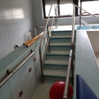 Scale e corrimano metallici all'interno di una piscina pubblica, pareti chiare e vista parziale su gradini e accessi, illuminazione naturale.