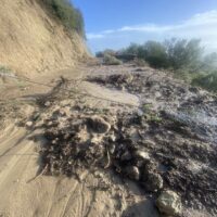 Sentiero sterrato tra scoscese scogliere e sassi, con vista mare in lontananza e vegetazione sparsа lungo i bordi
